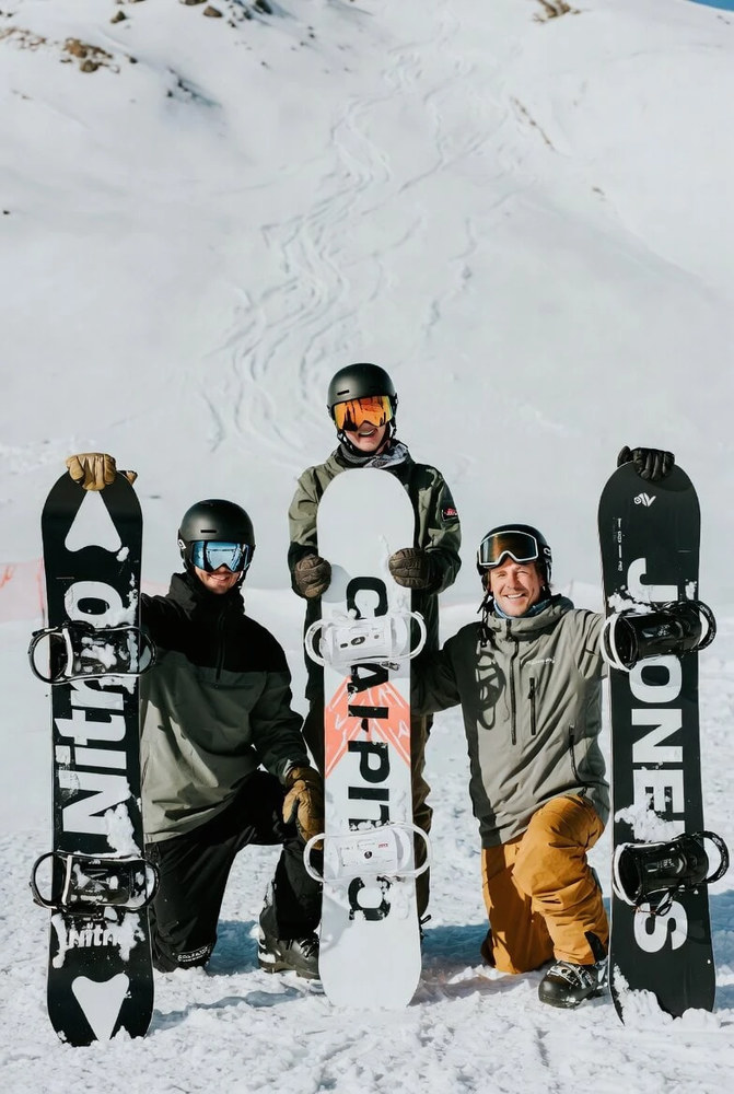 Троє друзів позують біля хвойного лісу на гірськолижному курорті: шоломи, маски, дошки Nitro, Capita і Jones, легке морозне повітря і ясне небо.