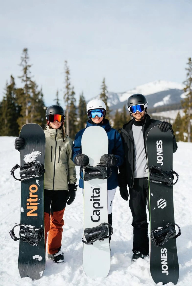 Три райдери відпочивають на сонячному схилі: двоє присіли в сніг, третій стоїть по центру, поруч сноуборди Nitro, Capita і Jones; позаду біла стіна.