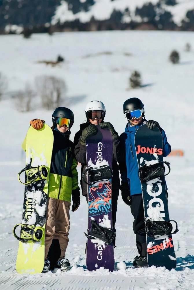 Три сноубордистки посміхаються на засніженій галявині біля лісу: сірі та бірюзові анораки, яскраві окуляри, дошки Nitro, Capita та Jones у пухнастому снігу.